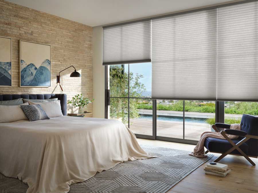 Alustra® Duette® Honeycomb Shades covering floor to ceiling black trim windows. The back wall has a bed on it with modern picture frames over the bed and a side lamp.