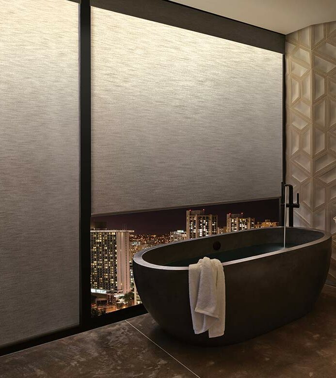a modern bathroom with a tub next to large windows covered by aura illuminated shades for privacy with warm light