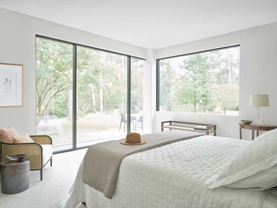 Floor to ceiling windows on two walls in an all white bedroom that overlooks a luscious backyard