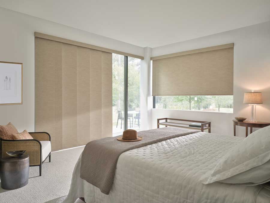 Floor to ceiling windows on two walls in an all white bedroom that overlooks a luscious backyard that has beige Skyline® Gliding Panel Track Blinds with Designer Roller Shades on the windows